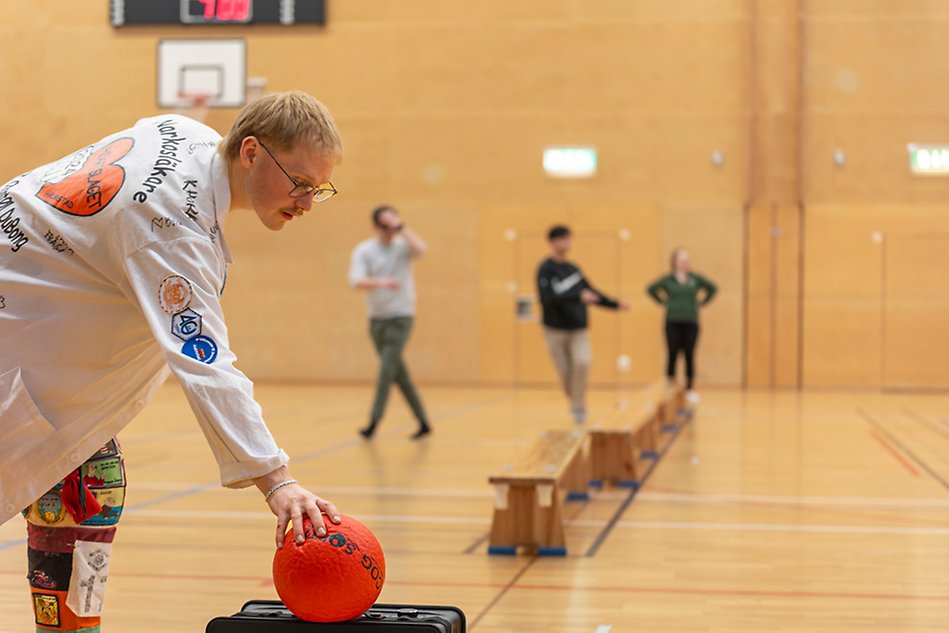 En person i en kraftigt dekorerad läkarrock sträcker sig efter en röd boll. I bakgrunden syns en idrottshall och några människor. Foto.