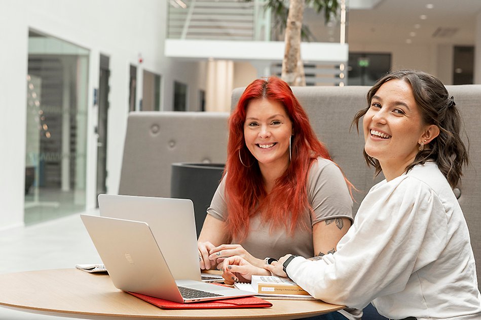 Two girls sitting with their laptops in a sofa, by a table. Both of them are smiling and looking in the camera. Photo.