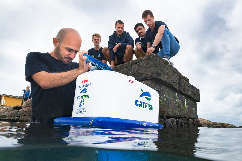 A group of people standing on a pier looking down at another person standing in water working with a drone. Photo.