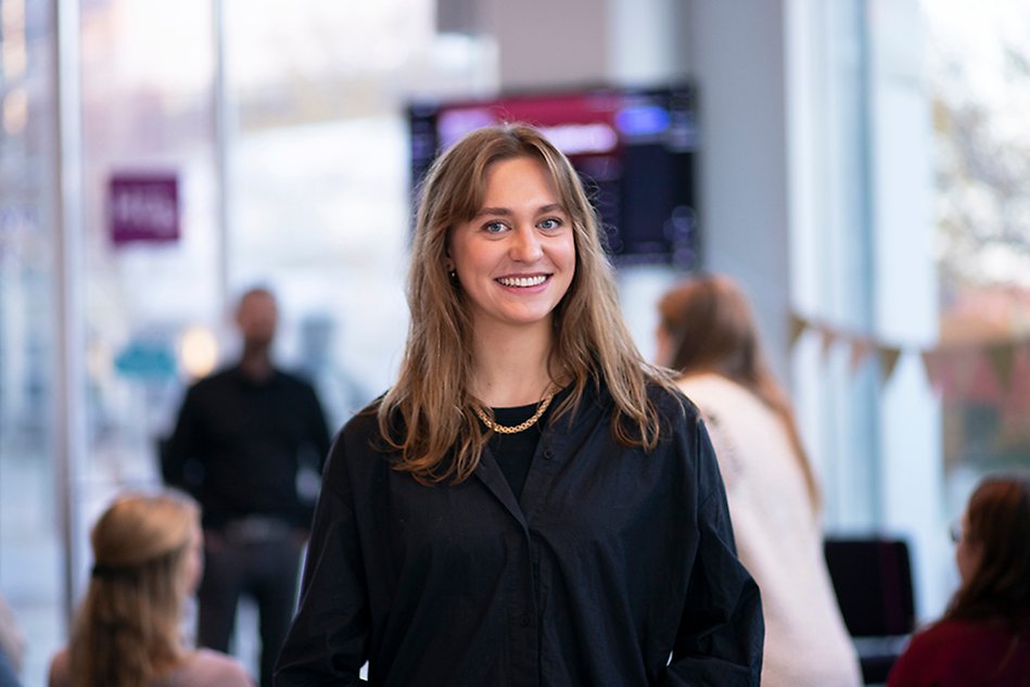 Leende kvinna med lugg och mellanlångt hår syns i förgrunden, i bakgrunden kan man ana en skärm på en vägg och tre personer i oskärpa. Foto.