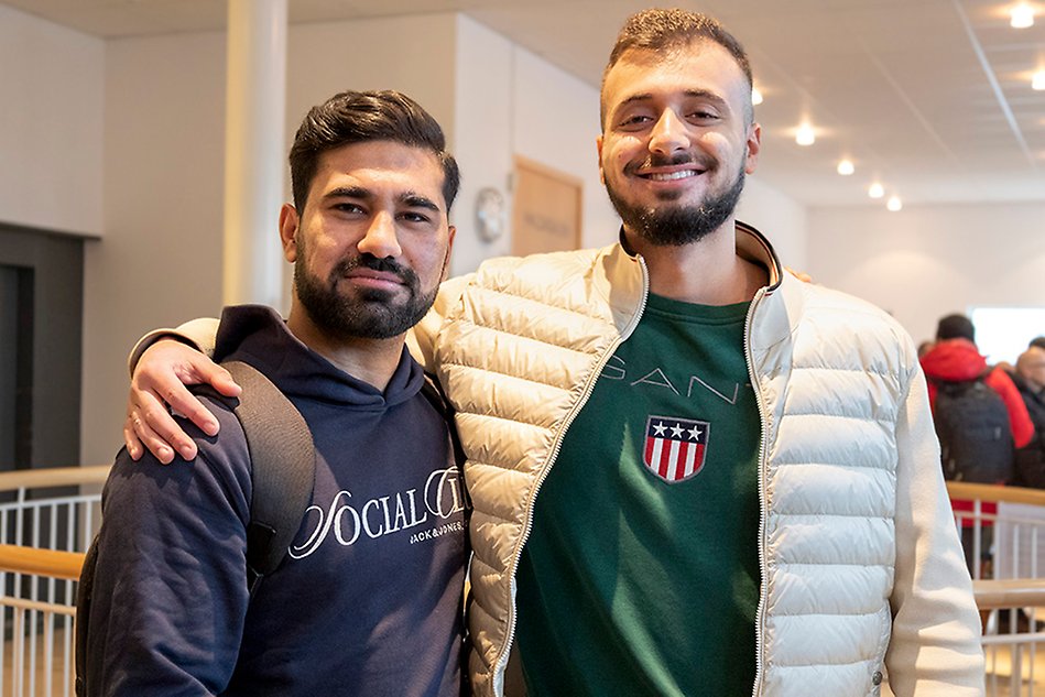 Two men are standing next to each other smiling, with the arm around one another. The man to the left wears a dark hoodie and the man to the right a bright jacket.