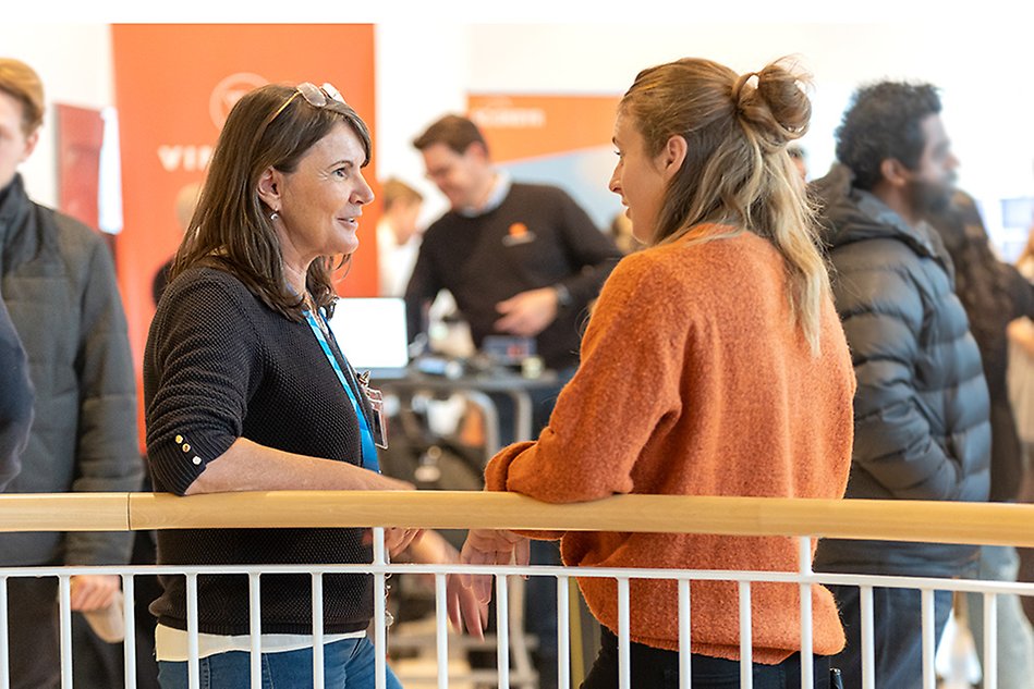 Two women are standing turned against each other and has a conversation. In the background there is a crowd of people. The photo is taken from the side.