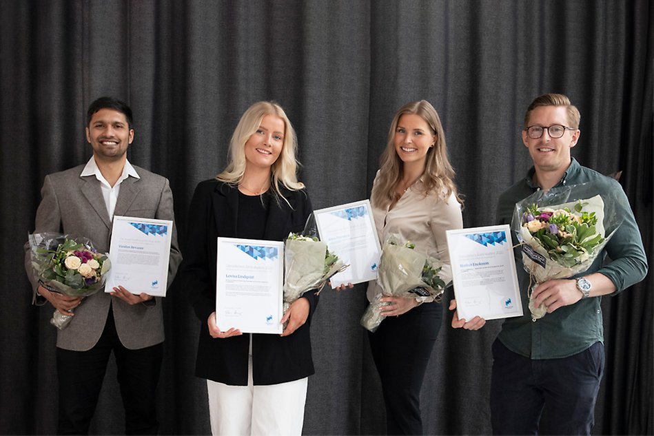 Four smiling people standing next to each other, each one of the is holding a diploma and flowers. Photo.