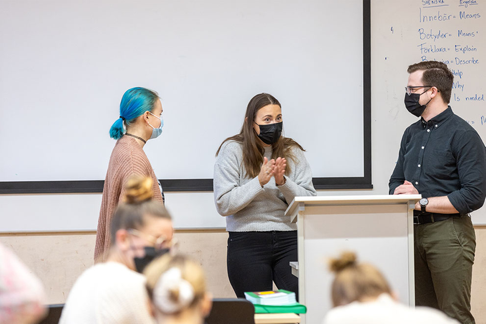 Tre personer som står bakom en kateder och håller tillsammans en lektion i svenska som andraspråk för studenter som sitter framför dem. På väggen hänger en whiteboard men svenska glosor skrivna. 