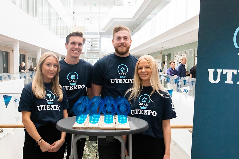 Fyra studenter poserar i t-shirt. Framför dem står ett bord fyllt med Utexpostatyetter. Foto.