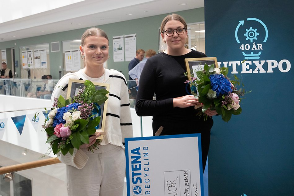 Two women in tied hair. The first one wearing black clothes and glasses, while the other one is wearing cream-coloured clothes. Both of them are holding diplomas, flower bouquets and a big check.