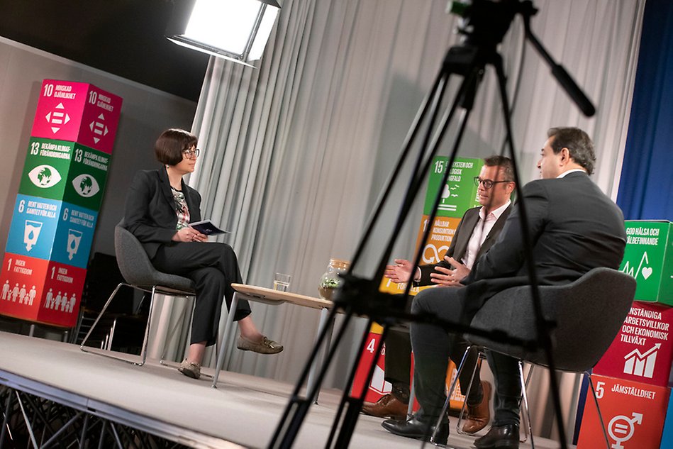 Three people on a stage. A camera tripod in the foreground.