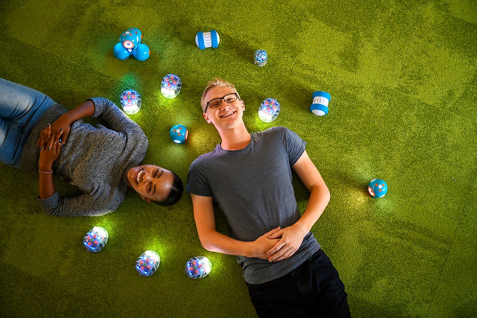 En man och en kvinna ligger på en grön matta och ler, runt dem små lysande robotar som liknar nyckelpigor eller skalbaggar