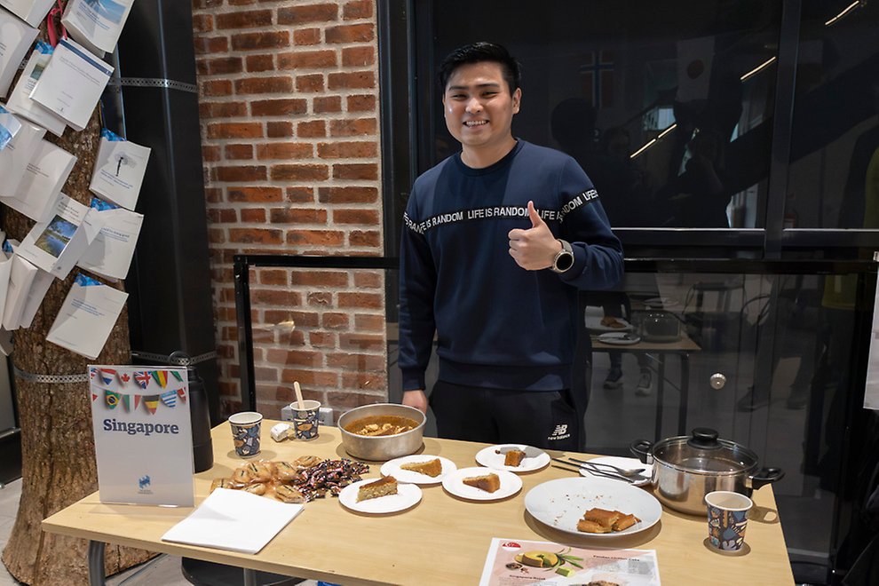 A person gives a thumbs-up from behind a table laden with plates of cakes. 