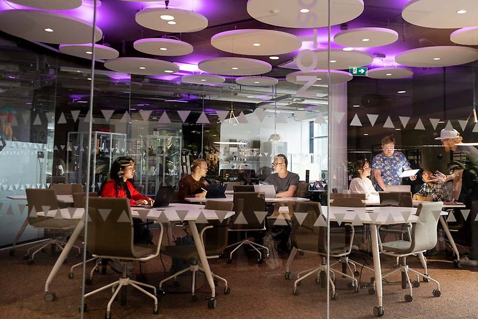 Through a window with stickers on, you see seven people around a table, five sitting, two standing. They seem to be talking to each other in two different groups. Photo