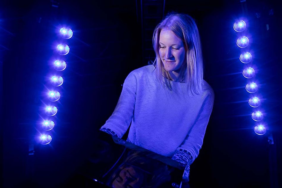 Woman looking down at a screen she is holding. The background is dark, black and deep blue. On each side of the woman shines a vertical row of lamps reminiscent of light bulbs. Photo
