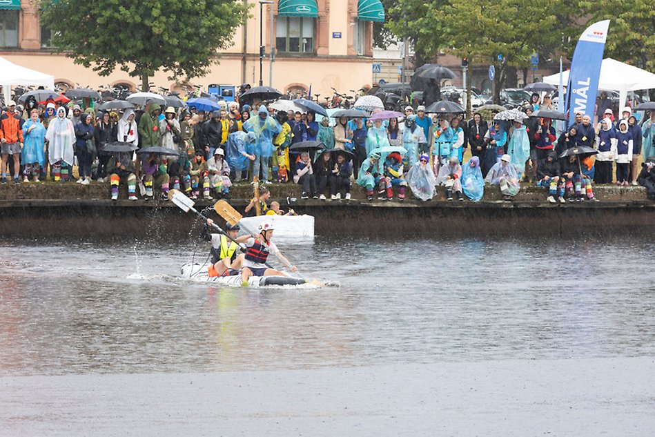 Två personer sitter på en avlång båt i en flod i stadsmiljö och paddlar. I bakgruden på land står en folkmassa med regnkläder och med paraplyer. Foto.