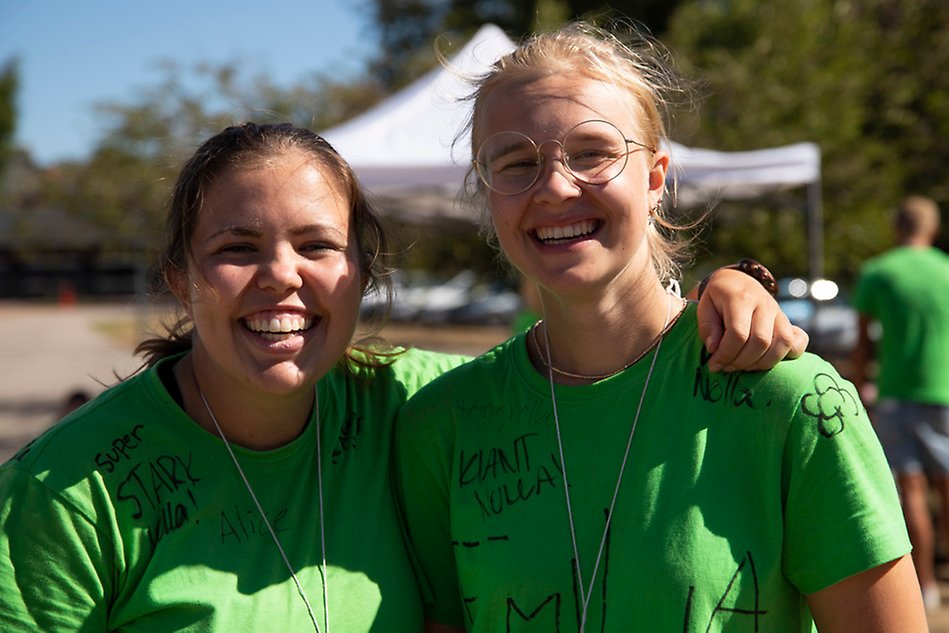 Två kvinnor i gröna t-shirts står tätt bredvid varandra och ler. Kvinnan till vänster i bilden har vänsterarmen om axlarna på kvinnan till höger. Foto.