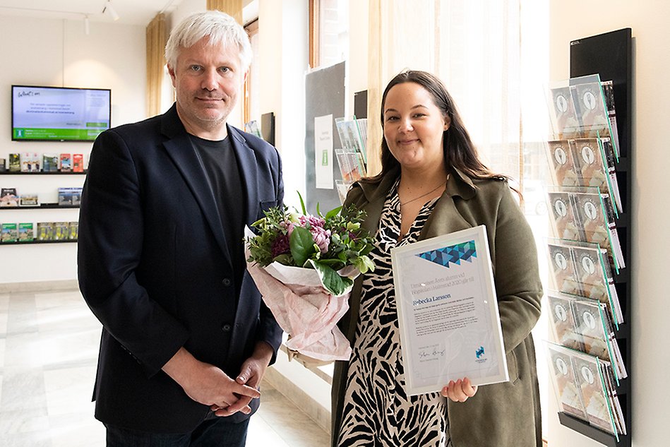 Två leende personer står bredvid varandra, en av dem håller ett diplom och blommor.