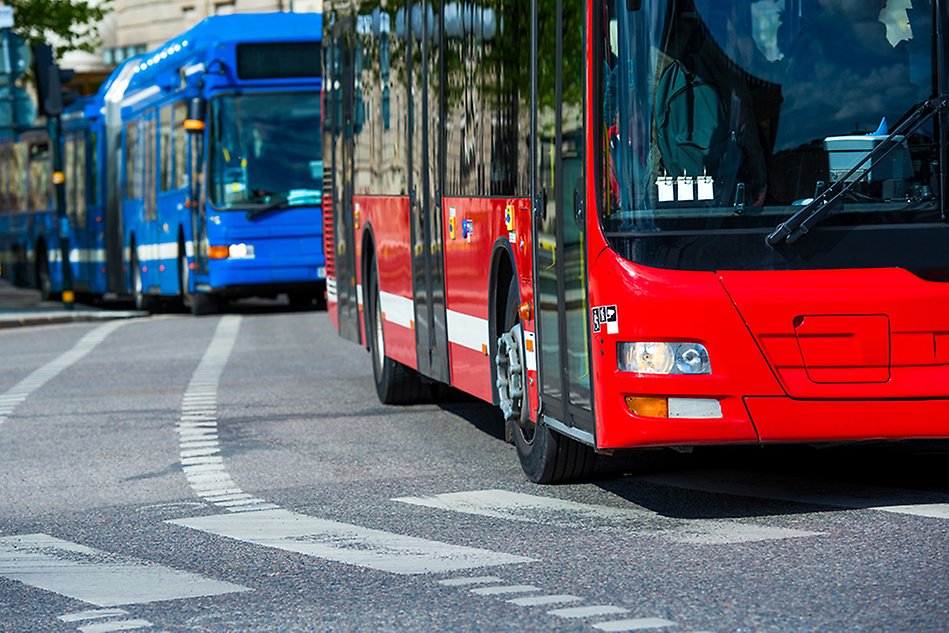 Red bus driving on a city street, followed by a blue bus. Photo.