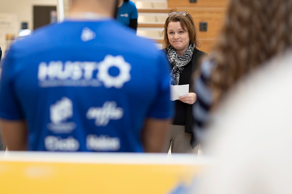 The foreground of the image shows the back of a man wearing a blue sweater. In the background, a woman with brown hair is smiling and looking ahead.