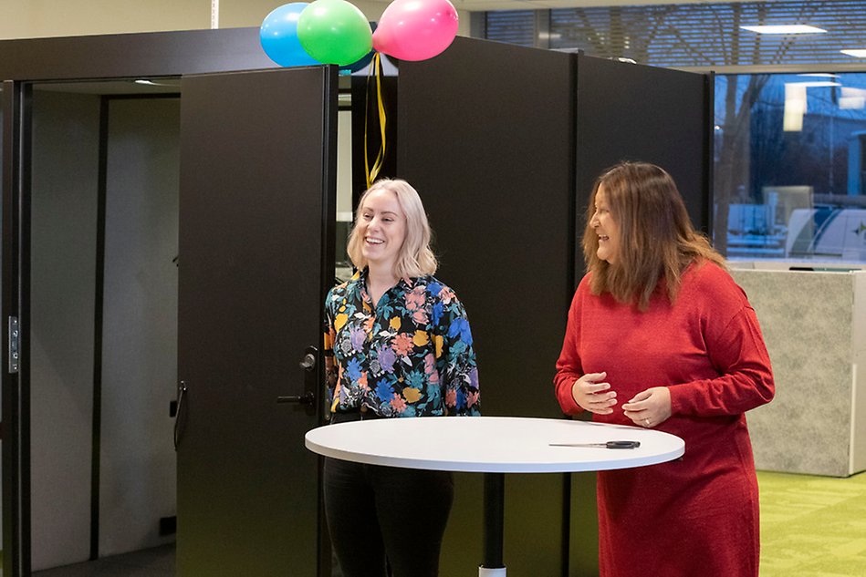 En blond och en mörkhårig kvinna står framför en svart liten byggnad inomhus, med ballonger fästa på.