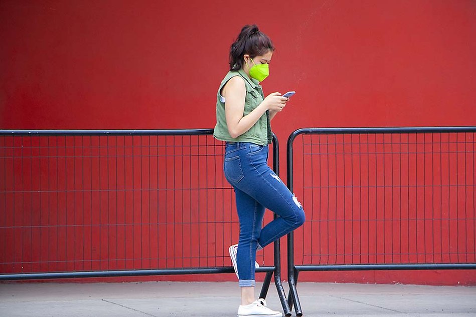 A woman leaning on a fence wearing a face mask and looking at a smartphone. Photo.