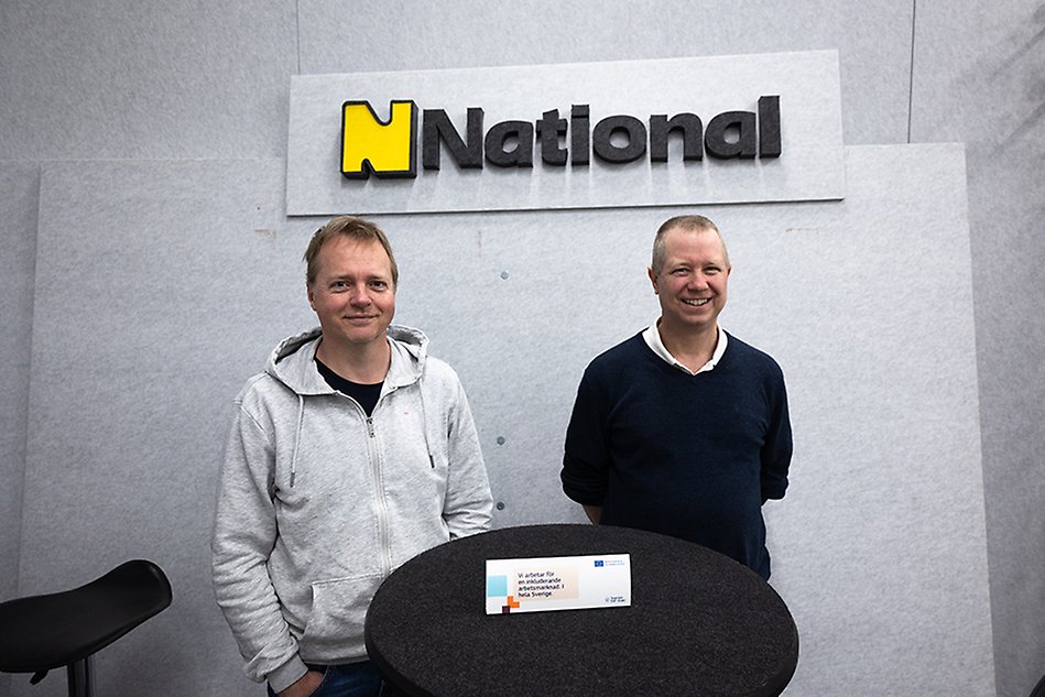 Två män står framför en logotyp där det står ”National”. Foto.