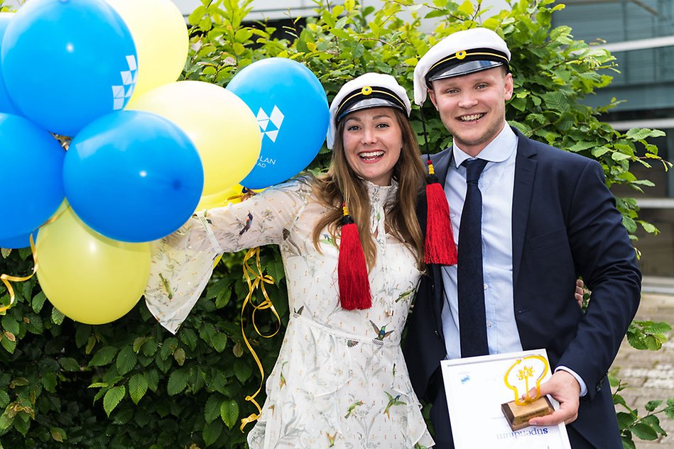 Två glada personer med finkläder och studentmössor. De håller i diplom. Till vänster gula och blå ballonger. Fotografi.