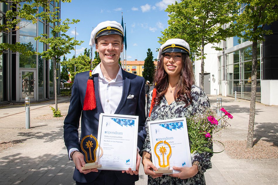 Två finklädda studenter står utomhus på campus och håller sina diplom och en statyett. De ler mot kameran. Foto.