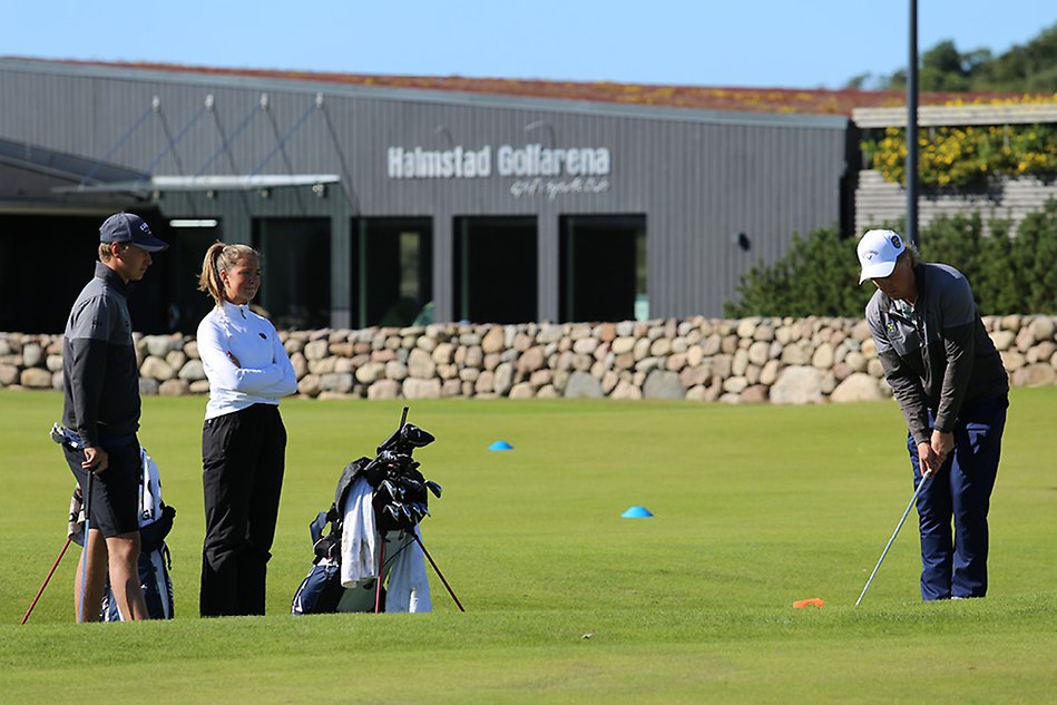 Två golfstudenter observerar sin tränare demonstrera hur man utför en teknik på en golfbana. Foto.
