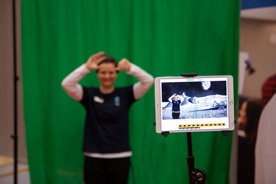 Grön skärm eller så kallad green screen, person I blå tshirt står framför den och på en läsplatta syns samma person på månen
