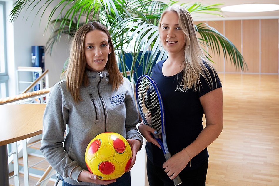 Two female students pose beside a green plant. One is holding a football in her hand and and the other is holding a racket. Photo.