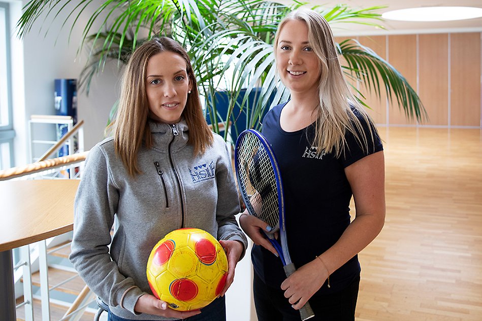 Två tjejer står bredvid varandra framför en palmliknande växt. Den ena håller en fotboll och den andra håller i ett racket. Foto.