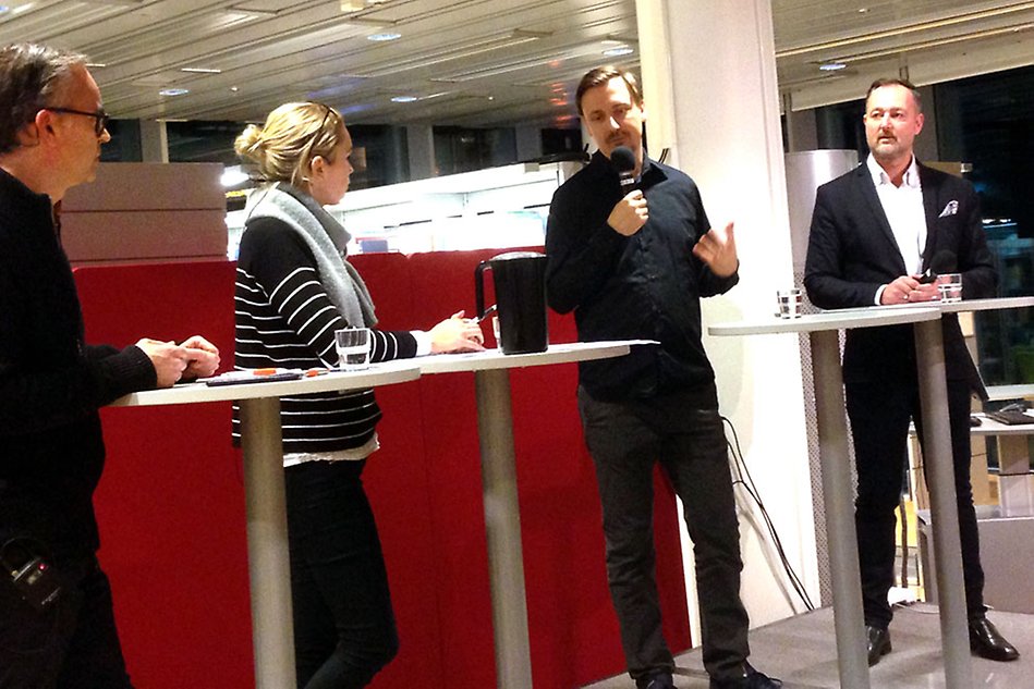 Fyra personer på en liten scen, en man pratar i mikrofonen. Röd bakgrund. Foto.