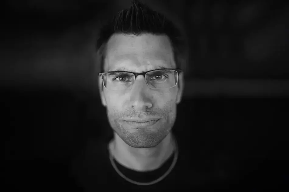 A person stands in a dark room, only the face is showing. The person has glasses. Photo.