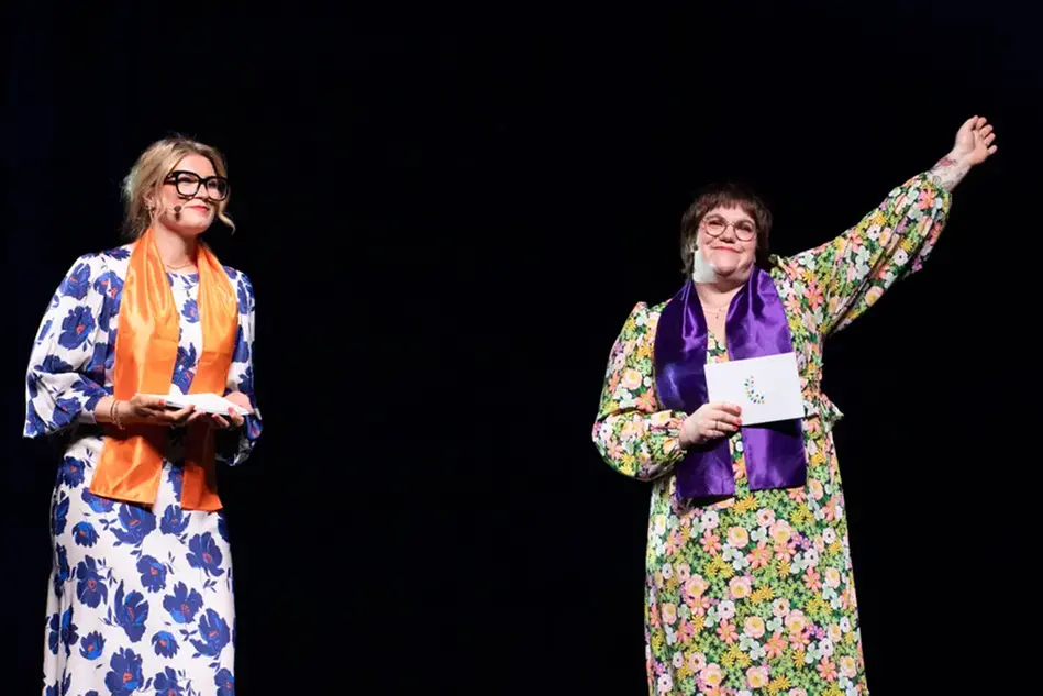 Two young women in colorful dresses stand on a stage and smile. Photo.