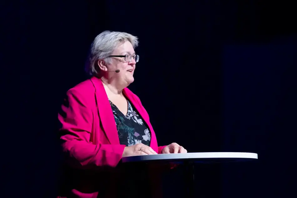 A woman with short blond hair and glasses stands at a podium. Photo.