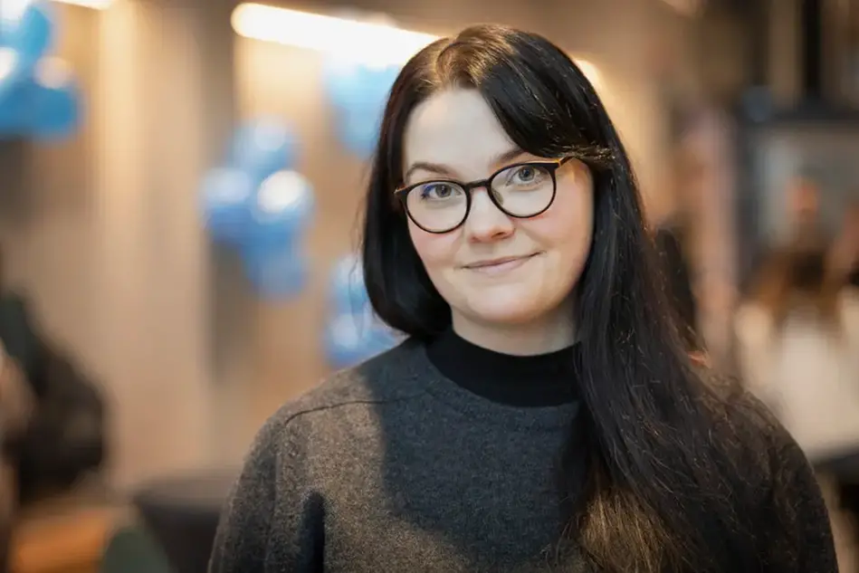 Kvinna i mörkt långt hår och glasögon ler mot kameran. I bakgrunden hänger ballonger. Foto.