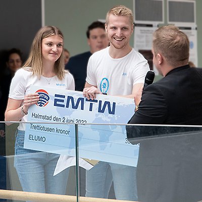 Två personer iklädda vit t-shirt och jeans håller i ett stort kort där det står "EMTW 30000 kronor ELUMO". De tittar på en person med en mikrofon som står i förgrunden. Foto.