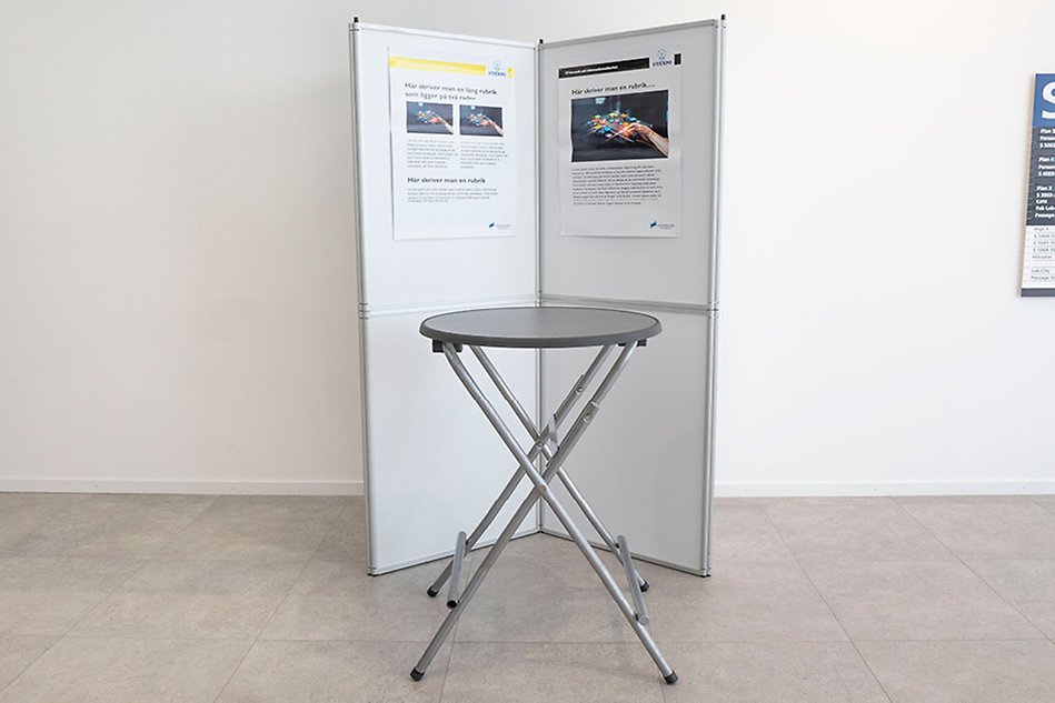 Two folding walls with posters are placed in front of a white wall. In front of them is a round standing table. Photo.