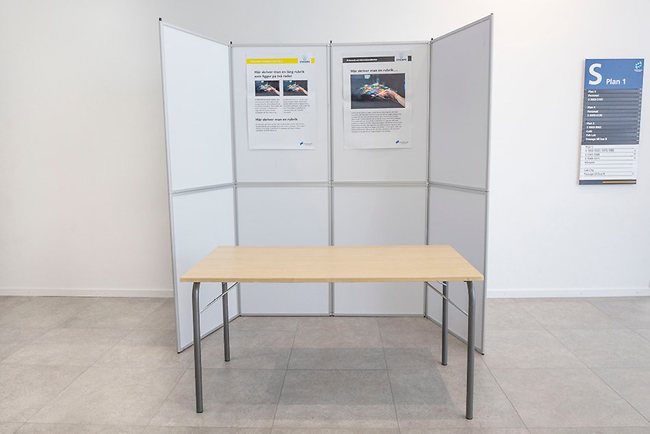 Four folding walls with two posters are placed in front of a white wall. In front of them is a low larger table. Photo.