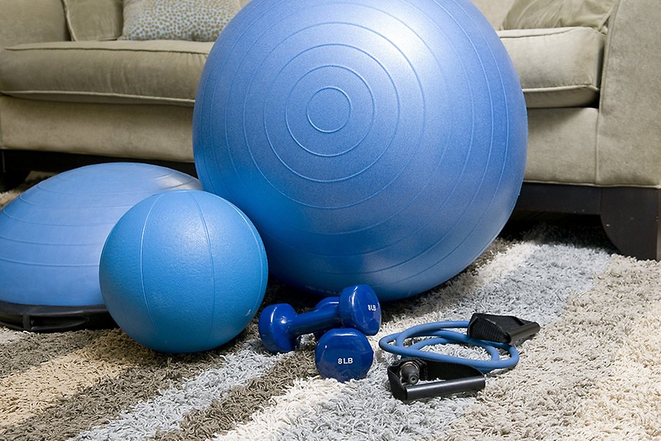 Träningsmaterial, som balansboll, träningsband och vikter, ligger på en matta i ett vardagsrum. Foto.