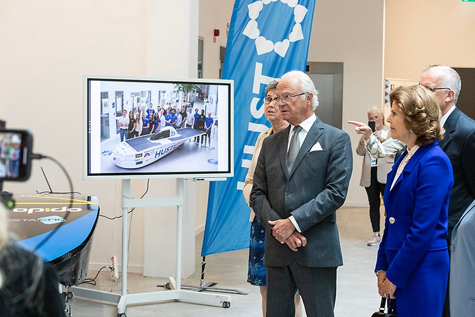 A group of people stand on the right and look to the left. In the background a screen showing a picture of a number of people standing around a car. Photo