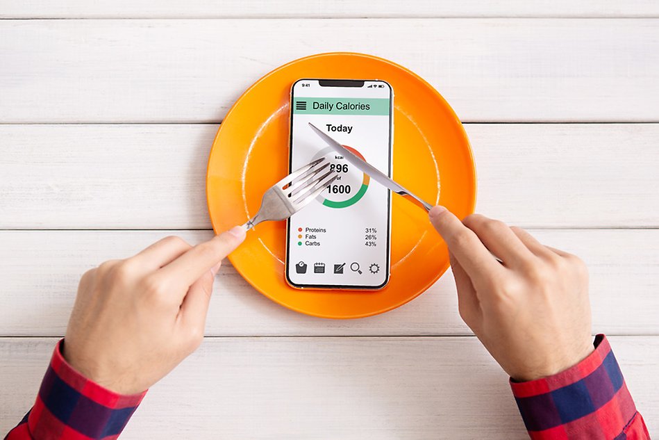 Yellow plate with smartphone on it showing daily calorie intake, two hands holdiing cutlery above the plate. Photo.