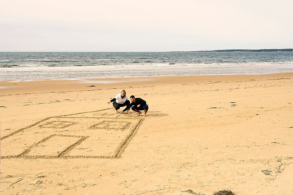 Två personer ritar ett stort hus i sanden på en strand. I bakgrunden syns havet. Foto. 