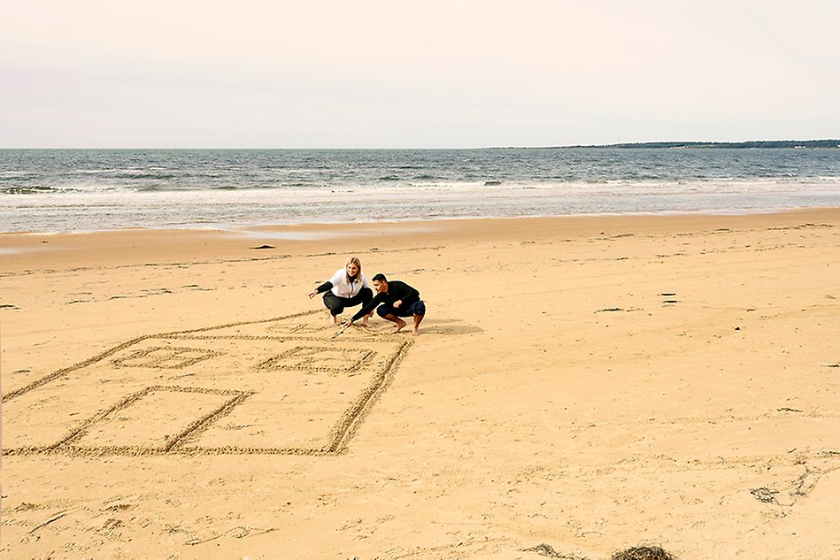 Två personer ritar ett stort hus i sanden på en strand. I bakgrunden syns havet. Foto.