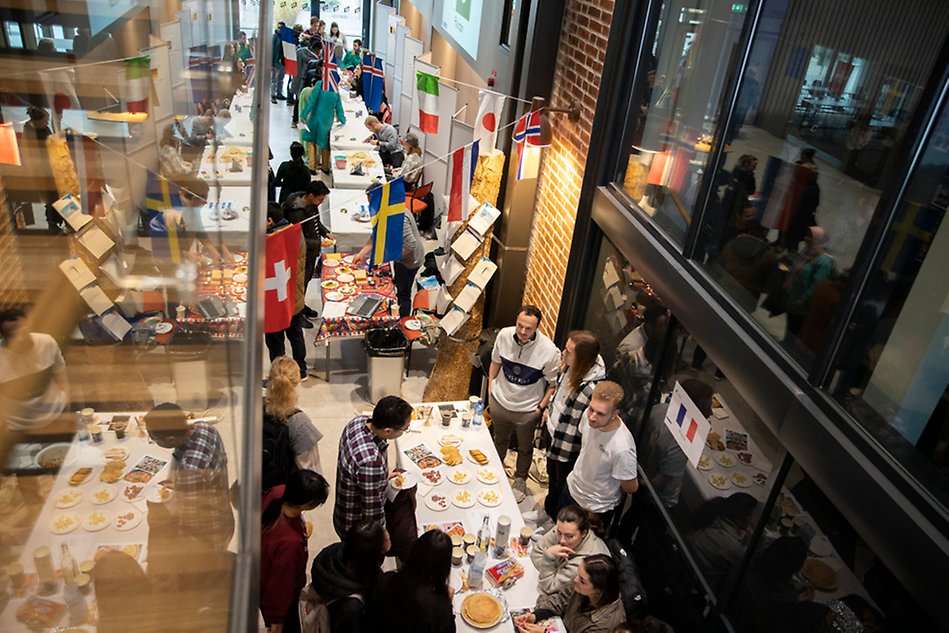 Photo from above. Tables line the walls with many people interacting. Flags are hanging from the ceiling and there is food on the tables.