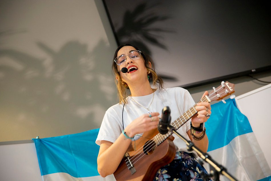 Person har mikrofon på sig och sjunger och spelar på en ukulele. En vit och blå flagga syns i bakgrunden. Foto.