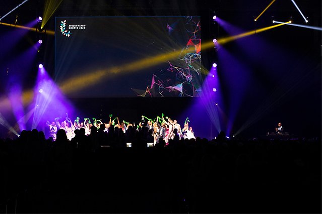 A crowd of young people on a stage that are waiving with colorful shawls.