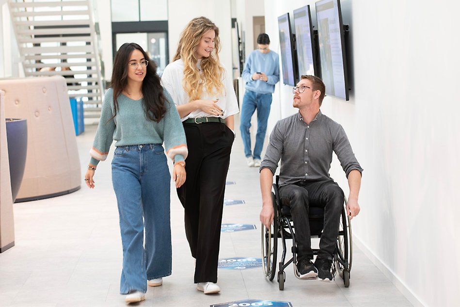 Three young people in a corridor. One person is in a wheelchair. In the background is another person. Photo