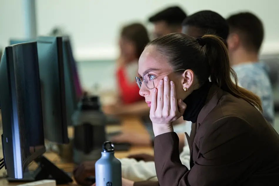 A young woman is sitting at a computer screen. Several people are visible in the background. Photo.