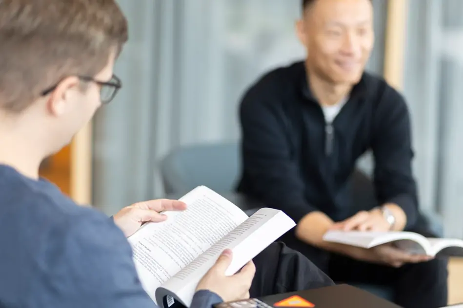 A student reads a book. Another student is visible in the background. Photo.