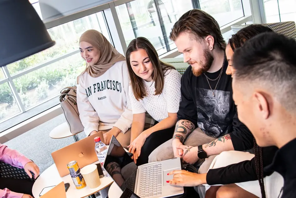 En grupp studenter sitter vid två mindre bord och pratar med varandra och ler. På bordet har de bärbarta datorer. Foto.