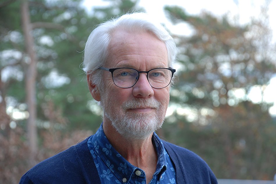 A man with white hair and black glasses standing outside looking into the camera. Photo.